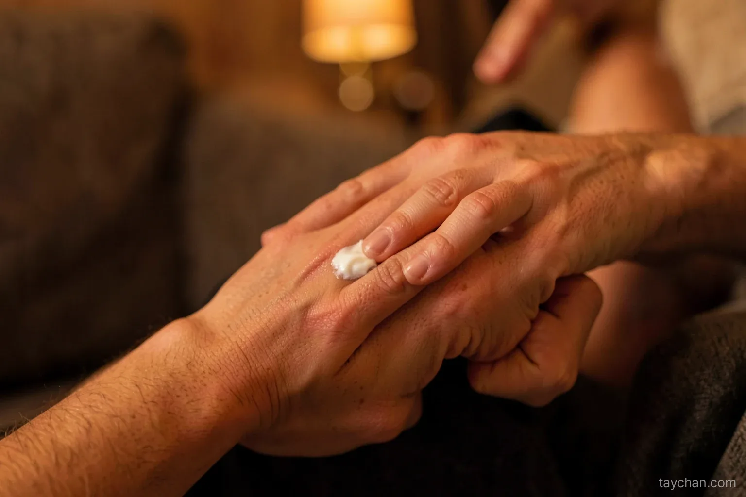 Close-up of a person rubbing a little unscented cream into the back of one hand with the opposite thumb, seated on a sofa in evening lamp light, warm amber tones, face not shown