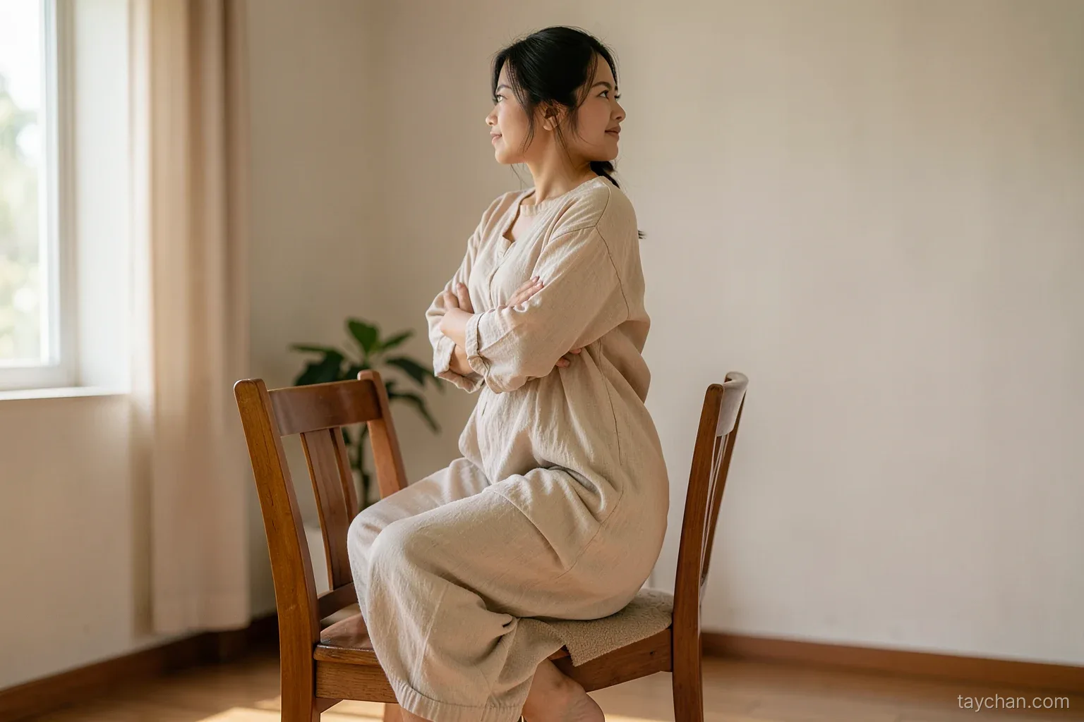 A Vietnamese woman in loose home clothes rising from a sturdy wooden chair with arms crossed over her chest, plain living room, morning daylight, side view, face partly turned away