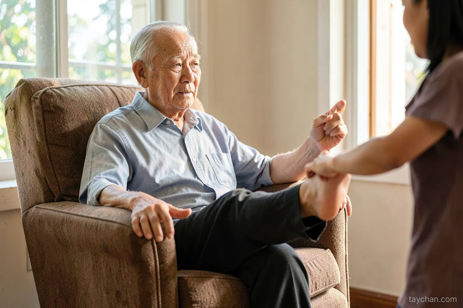 An elderly Vietnamese man seated in a sturdy armchair by a sunny window, lifting one foot and pointing his toes, his daughter's hand resting on the chair back, faces out of frame